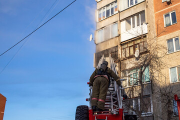 a firefighter extinguishes a balcony, in a high-rise building, from a telescopic ladder of a fire engine using a hydrant, against a blue sky