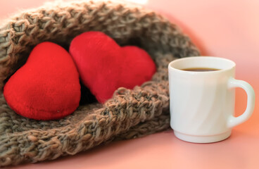 White mug with black coffee on the background of two blurry red plush hearts lying in a knitted scarf on a pink background. A pair of red hearts. Mutual love. The Concept Of Valentine's Day.