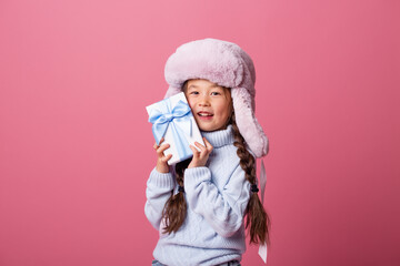 Obraz na płótnie Canvas Cute little Asian girl in a winter hat and sweater holds a gift box. Christmas concept, text space