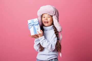 Cute little Asian girl in a winter hat and sweater holds a gift box. Christmas concept, text space