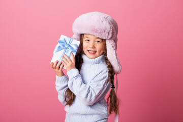 Cute little Asian girl in a winter hat and sweater holds a gift box. Christmas concept, text space