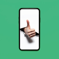 Close-up of human hand showing thumbs up through white paper hole on screen of smartphone; isolated on background of green color.