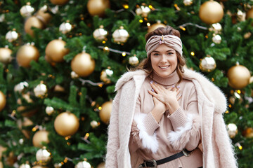 Outdoor portrait of young beautiful happy smiling girl posing on street against background of decorated fir  tree.   Christmas, new year, winter holidays concept. City lifestyle.