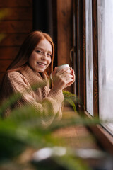 Fototapeta premium Portrait of charming smiling young woman standing near window with cup hot coffee and looking at camera. Side view of happy charming red-haired lady having rest at home.