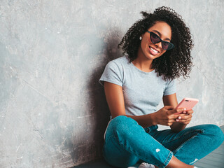 Beautiful black woman with afro curls hairstyle.Smiling model in white trendy jeans and sunglasses. Sexy carefree female sitting on the floor near gray wall in studio. Looking at cellphone screen