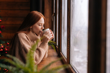 Side view of charming smiling young woman looking out of window and drinking coffee at home at Christmas time. Concept of home festive atmosphere.