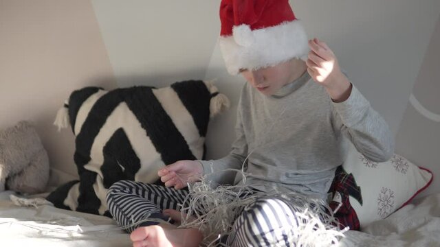 A child in the cap of the grandfather of frost untangles the New Year's garland for the Christmas tree.