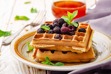 Closeup view at belgian waffles served with blackerries and mint leaf on white wooden kitchen table with syrup aside
