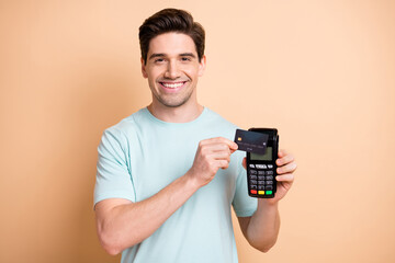 Close-up portrait of nice cheerful guy holding in hand terminal using bank card finance transaction isolated over beige pastel color background - obrazy, fototapety, plakaty