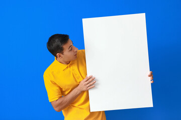 Surprised young man with blank poster on color background