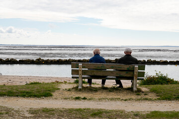 senior person retired elderly couple sitting on wooden bench looking ocean atlantic seaside in low tide