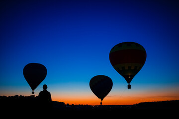 Three Amazing Colorful Air Balloons Levitating Over the Ground Outdoors With A human Silhouette Against Blue Skies At Twilight.