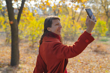 A beautiful young woman makes a video call on a smartphone, a woman stands on an alley in a Park in autumn, has a rest online-chat with friends, a student talks, a blogger runs a vlog blog