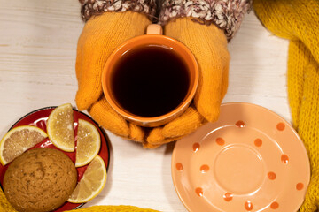 Two hands in orange gloves hold a cup of coffee. Nearby is a saucer of lemon and oatmeal cookies.