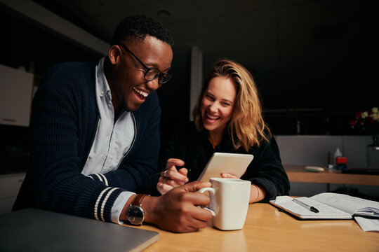 Happy businessman and businesswoman sitting together laughing while looking at digital tablet during break time at night - two colleagues smiling and working late together