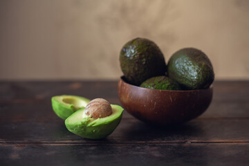 Whole avocado fruits in a bowl made from coconut shells and cut in half lie next to it on a dark wooden background