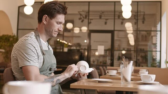 Waist up side view of middle-aged Caucasian male public catering worker wearing apron, sitting by table alone in restaurant hall, wiping cup with towel, then putting it down
