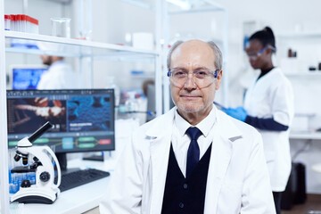 Senior scientist sitting at his workplace during medical experiments. Elderly scientist wearing lab coat working to develop a new medical vacine with african assistant in the background.