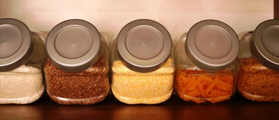 various gluten-free cereals and pasta are in glass containers with gray plastic lids on a brown shelf as a fragment of the kitchen interior