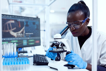 Virologist of african ethnicity sitting at her workplace neat test tubes looking through...