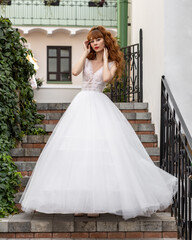 Beautiful bride in a big dress with red curly hair stands on the steps of the stairs against the background of the old city. Exterior fashion shot of fashion model in wedding dress
