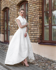 A luxurious bride in a white long wedding dress standing on the street of an ancient city against the background of a brick wall with windows of a building. Model posing for a photographer