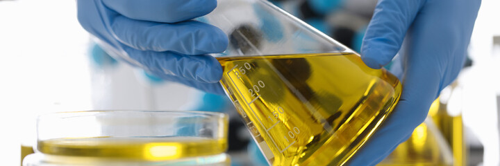 Hands in rubber gloves hold flask with yellow liquid in chemical laboratory closeup. Quality control in oil refining industry concept.