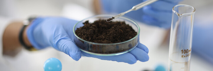 Glass cup with soil sample stands on hand in rubber glove in chemical laboratory closeup....