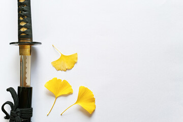 白い紙の背景の左に置いた抜きかけの日本刀と三枚の黄色いイチョウの葉。右にコピースペース。