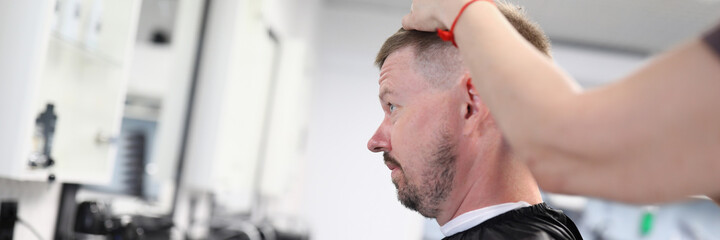 Obraz premium Young man sits in hairdressing chair and makes haircut in beauty salon portrait. Small business development hairdressing services concept.