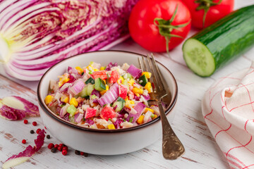 Delicious red Peking cabbage salad with tomatoes, cucumbers, corn, onions and crab sticks in a white plate on a wooden table. Light gourmet dinner. Selective focus