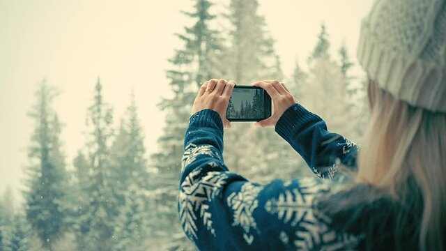 Woman traveler with her phone camera taking pictures beautiful winter snowy forest view. Fashion girl in wool sweater. Happy winter holidays. New Year's Eve. Christmas holiday. 4K video Slow motion.
