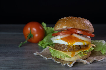 Burger, hamburger, cheeseburger, in bun with lettuce, tomato, onion, fried egg, delicious fast food, on gray wooden, black background.