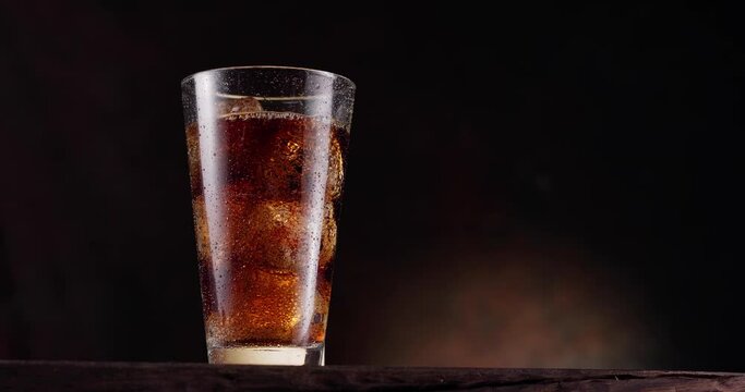 Glass of cola with ice stands on a wooden table with a black background. Gas bubbles actively rise in the glass. Slow motion, Blackmagic Ursa Pro G2.