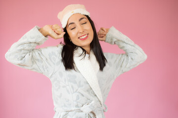 Relaxed joyful woman stretches hands after awakening, rejoices good day, enjoys pleasant sleep, wearing nightgown, has happy expression, poses over pink wall