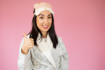 Young happy cheerful woman in sleeping clothes showing thumb up over pink background.