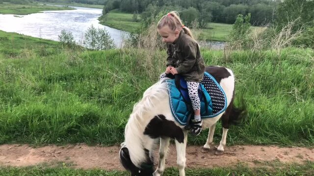 A beautiful little girl learns to ride a pony in the summer in a field by the river in Russia.