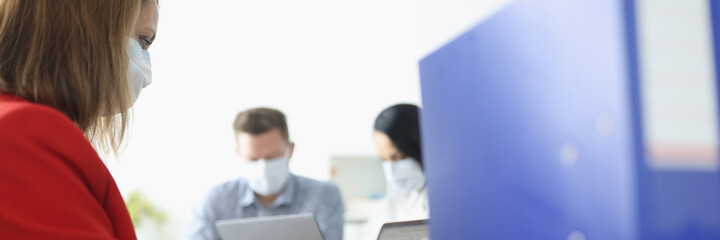 Young woman in business suit and masked face is typing on laptop keyboard in office with colleagues on background. Keeping distance in workplace during covid 19 pandemic concept.