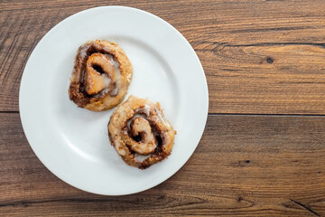 Hand made tasty cinnamon roll on a white plate and wooden table, top view. Pastry product.