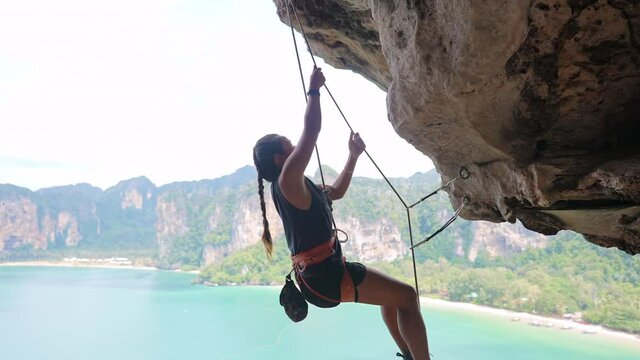 Confidence Asian woman climber with safety rope climbing high up on rocky mountain at tropical island in summer. Healthy and strong female enjoy active lifestyle and extreme sport in holiday vacation.