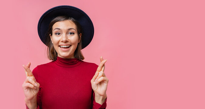 Nice cheerful woman wishes to do her best to keep her fingers crossed, has the best, wearing in  red jamper isolated on  pink background. People, body language concept. Copyspace.