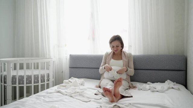 Happy pregnant woman looking at baby clothes with tenderness while sitting on sofa in domestic room. Caring future mother making purchases for unborn baby before childbirth. High quality 4k footage
