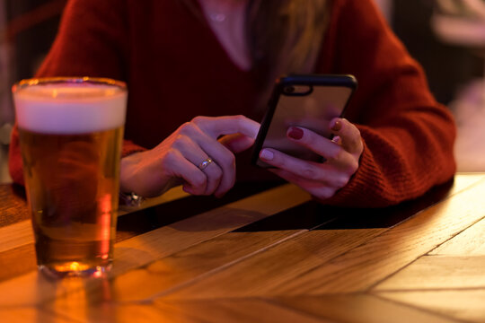 A young woman uses her mobile phone. She has painted nails and a ring. Next to her is a glass of beer on a wooden table