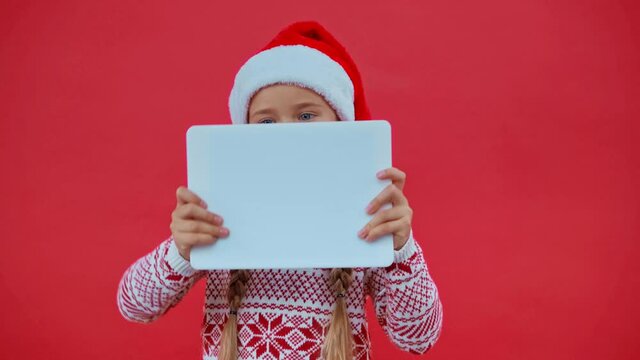 girl in santa hat and christmas sweater playing on digital tablet isolated on red