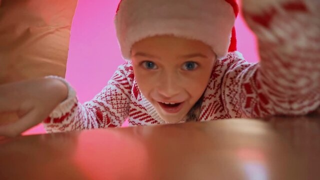 surprised girl in santa hat and christmas sweater looking in box isolated on pink