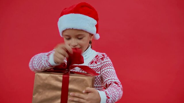 girl in santa hat and christmas sweater opening gift box isolated on red