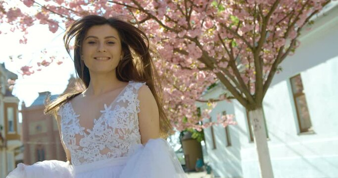 Beautiful smiling brunette with natural make-up is spinning round in the white pompous dress while walking along sunny street. 4k footage.
