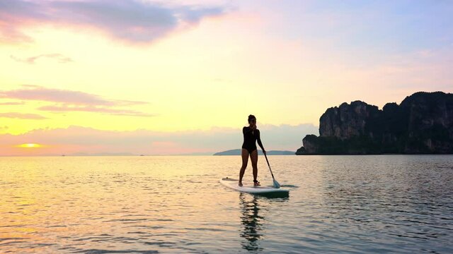 Silhouette of Confidence Asian woman standing on sup board with paddleboarding passing through beautiful sea and twilight sky at summer sunset. Female enjoy recreation water sport in holiday vacation