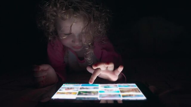 Close up of adorable curly-haired girl of four years old uses a tablet at night in bed.  Little girl scrolls through the photos on her tablet before bed.