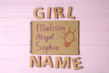Notebook with written different baby names and wooden letters on pink wooden background, flat lay